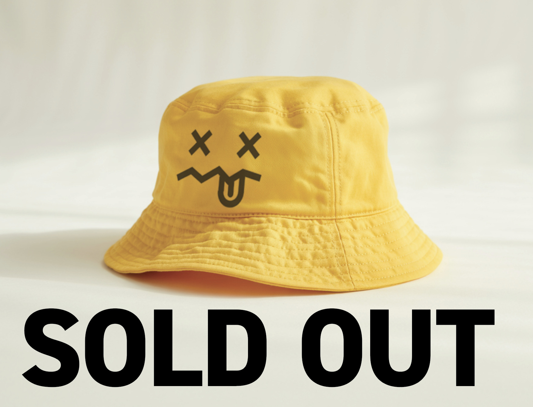 Yellow bucket hat with a cartoon face of X's for eyes, a wavy mouth, and a tongue sticking out.