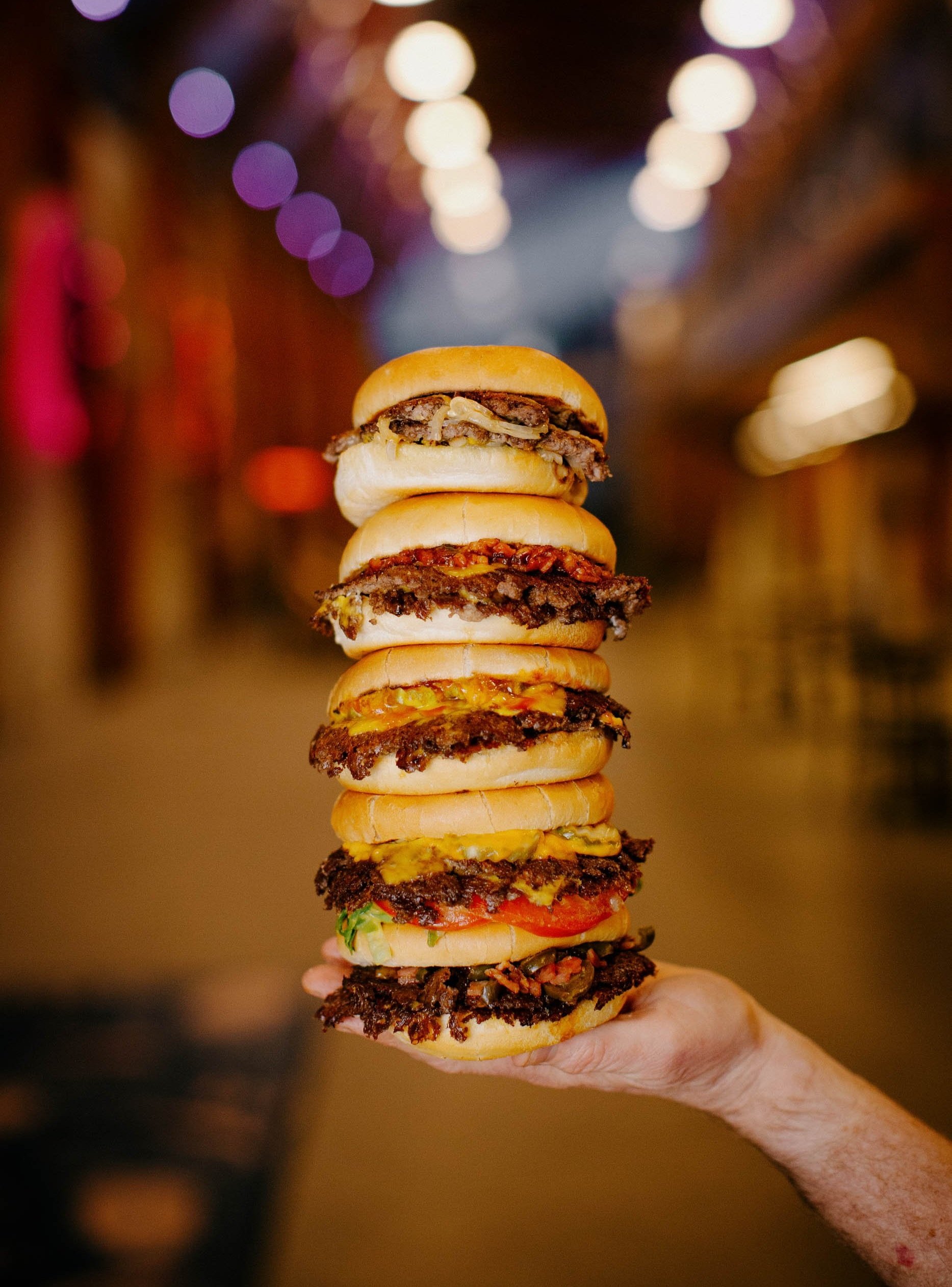 A tall stack of five smashburgers with various toppings in a dimly lit setting with colourful lights in the background.