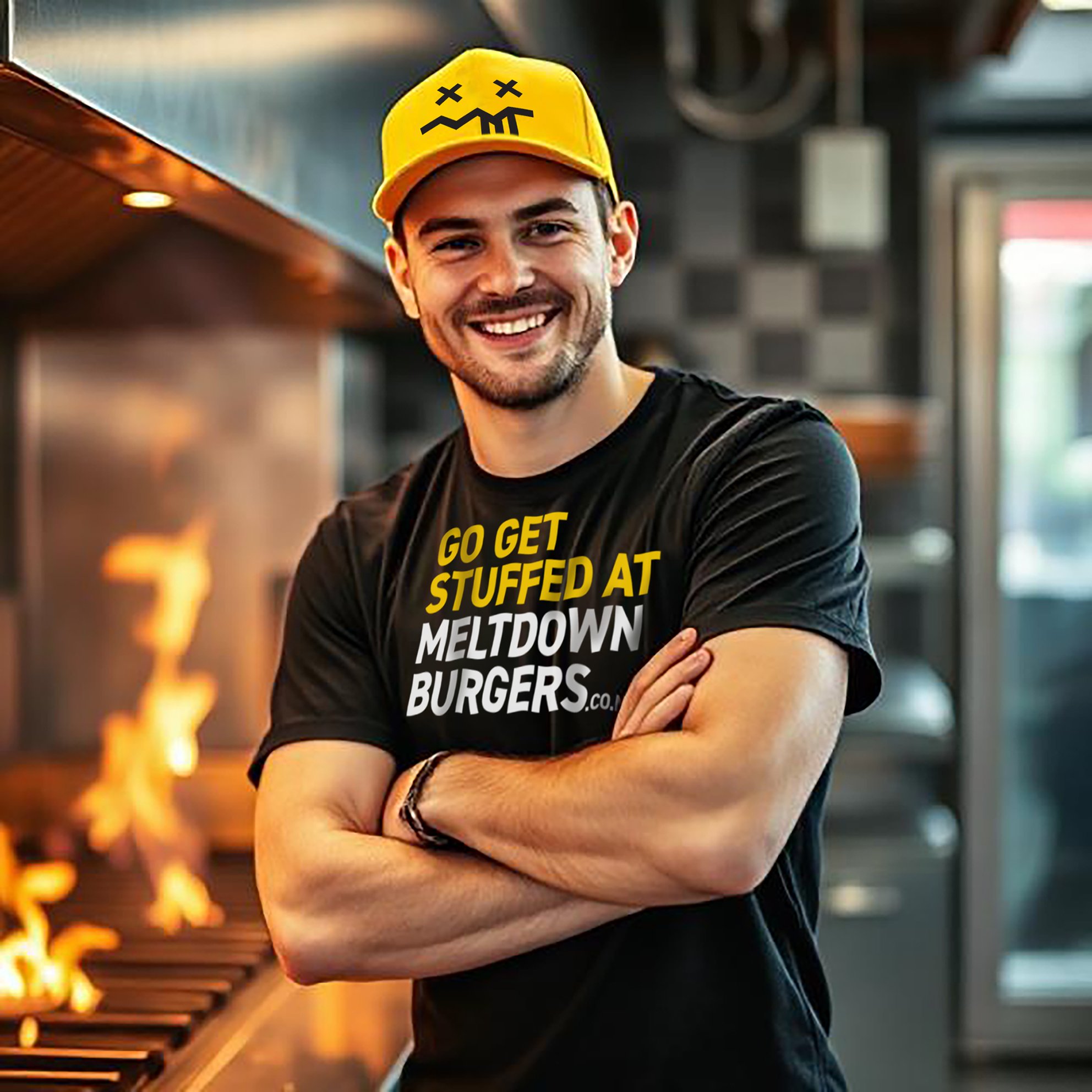Smiling man in a black T-shirt with yellow and white text and a yellow cap, standing in Meltdown Burgers restaurant kitchen near a grill with flames.